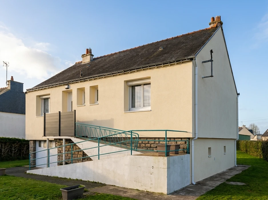 Maison individuelle de plain-pied surélevée, façade enduit lisse ton crème, toiture à deux pans en ardoise/tuile fibrociment grise, deux cheminées en briques